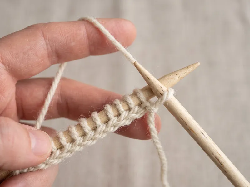 Close-up macro of long-tail cast-on: holding the yarn in a slingshot grip between thumb and index finger as the needle scoops the loop